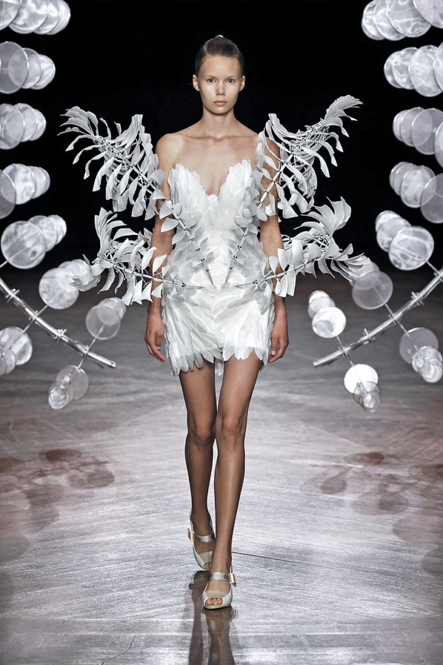A model walks through the Omniverse sculpture by Anthony Howe wearing a white dress with symmetrical metal bands of rotating silk leaves