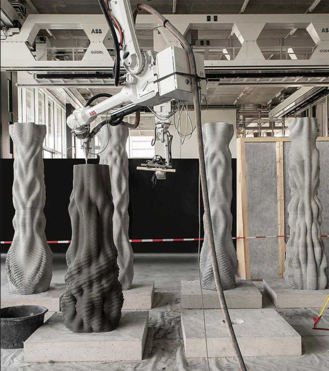 A robotic arm deposits concrete on a half-complete columns. Four completed columns stand behind it inside the fabrication space.