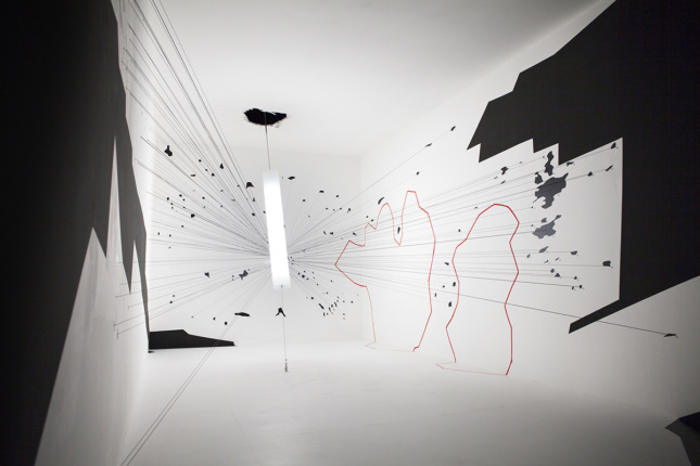A white room with black cutouts and splatter, as well as red outlines. A cylinder comes out of a hole in the ceiling to which lines are attached.