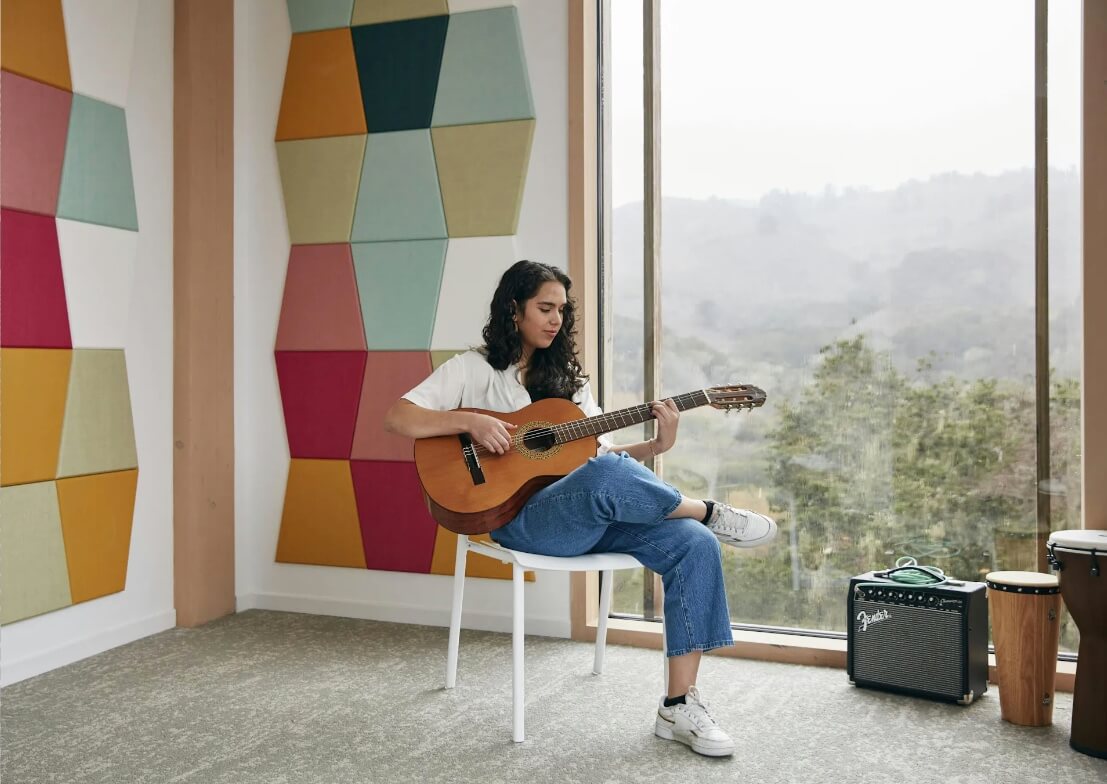 a girl plays in guitar in a brightly colored breakout room with large windows